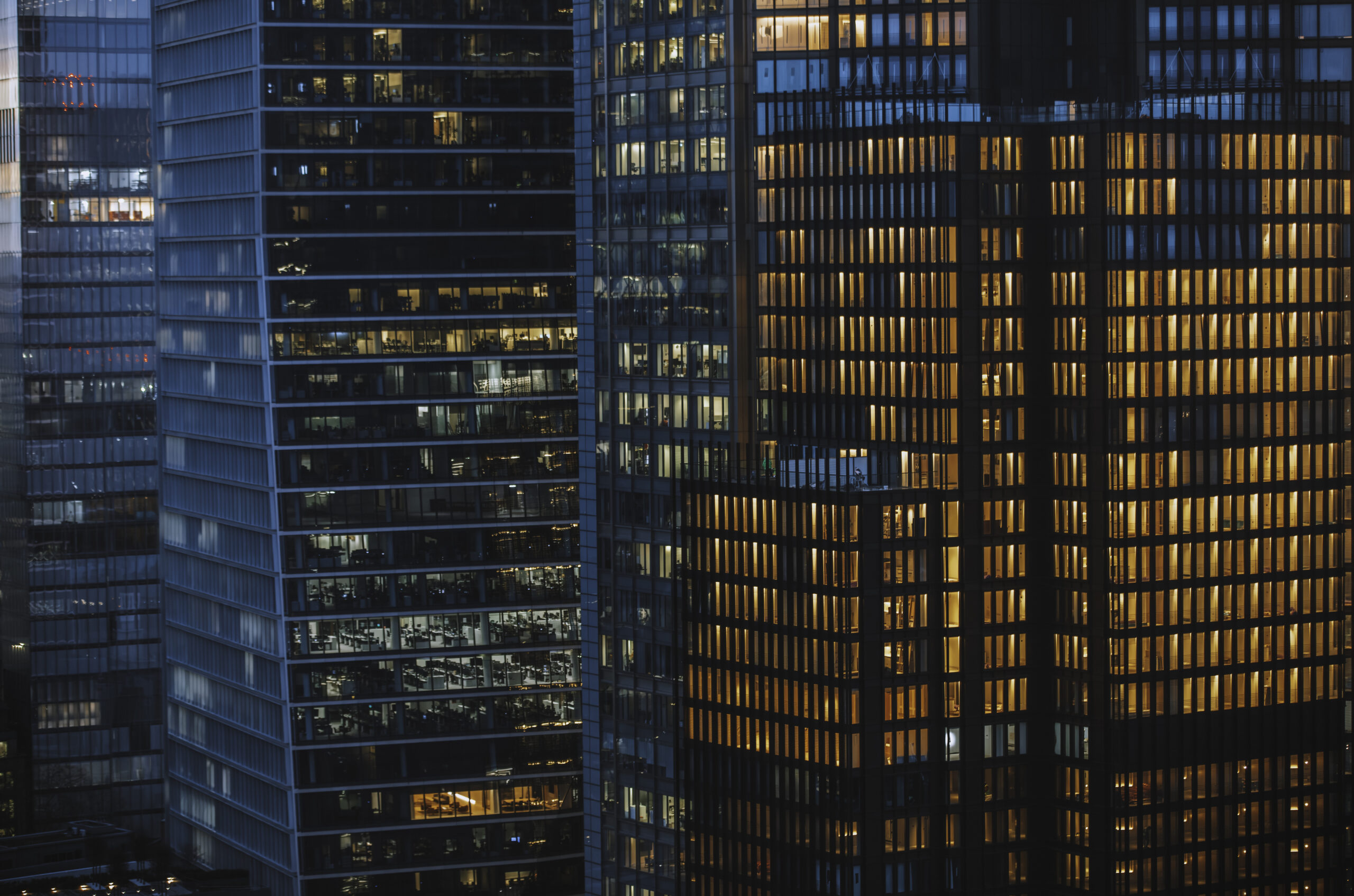 Close-up of illuminated glass skyscrapers in London’s financial district during twilight. The warm light glowing from the office windows creates a striking contrast with the dark reflective glass, showcasing the modern architectural design and the bustling corporate environment of the city